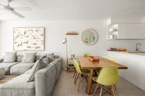A modern living room with a sofa, a dining table with chairs, a ceiling fan, a wall-mounted mirror, and a kitchen area with cabinets and a sink. - Headland Views 2 - Leafy Retreat (Noosa)
