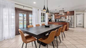 Uma cozinha moderna com eletrodomésticos de aço inoxidável, uma área de refeições com uma mesa grande e cadeiras, e vista para uma piscina através das portas de vidro.
