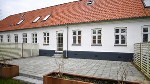 Ein Gebäude mit rotem Ziegeldach, weißen Wänden und zahlreichen Fenstern. Im Vordergrund befinden sich zwei Holz-Pflanzkästen mit Pflanzen.