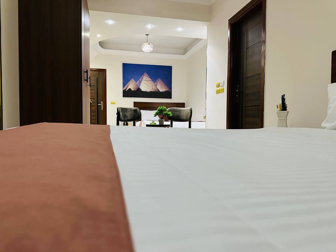 Une chambre d’hôtel avec un grand lit, un bureau avec une chaise, une petite table avec une plante et un tableau représentant des pyramides accroché au mur.