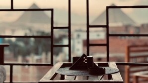 A view through a window with a geometric pattern, featuring a table with a coaster and a cup, and a cityscape with pyramids in the background. - MARQUIS PYRAMIDS HOTEL (Giza)