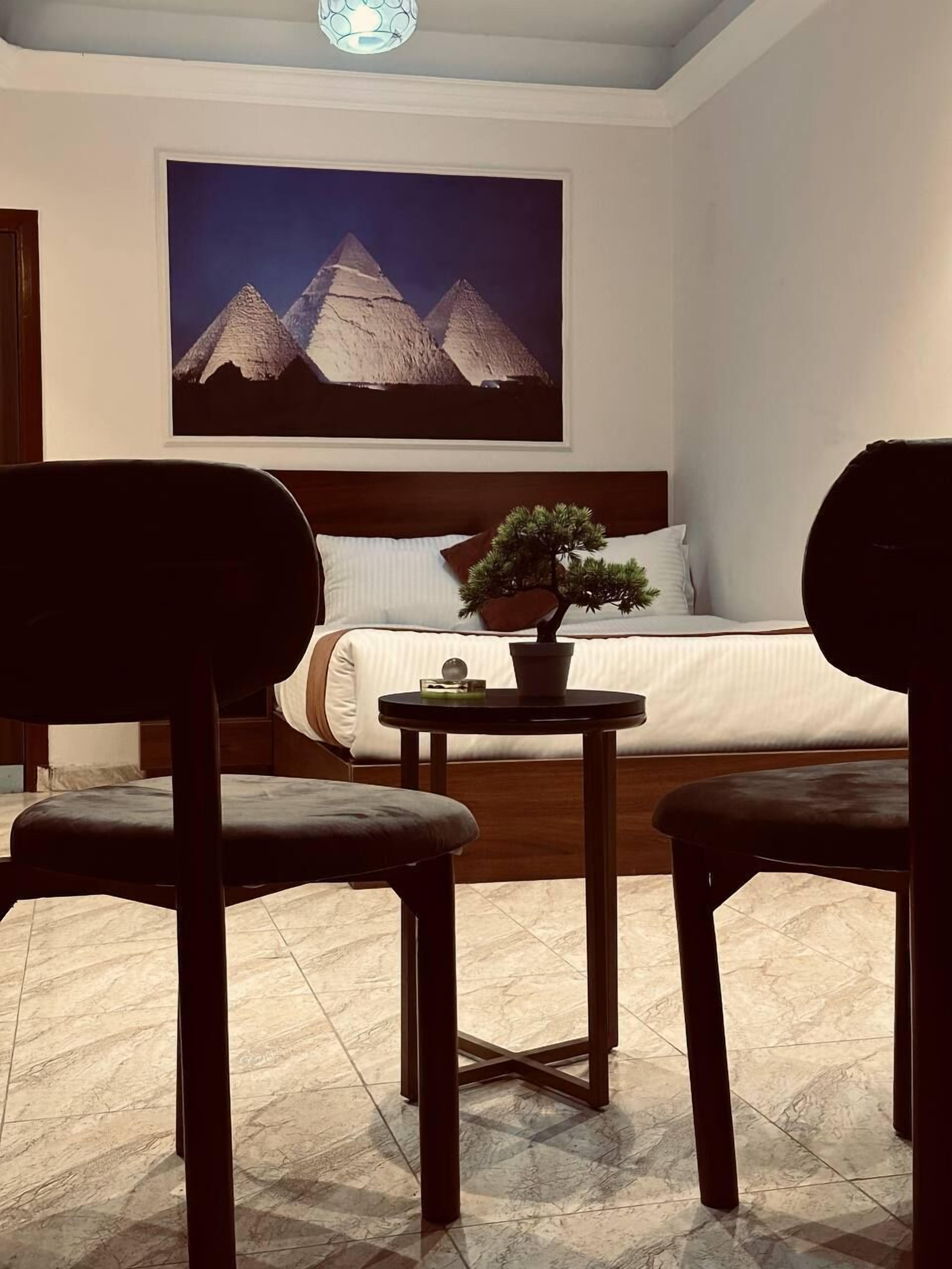 A hotel room with a bed, a nightstand, a potted plant, and a painting of pyramids.