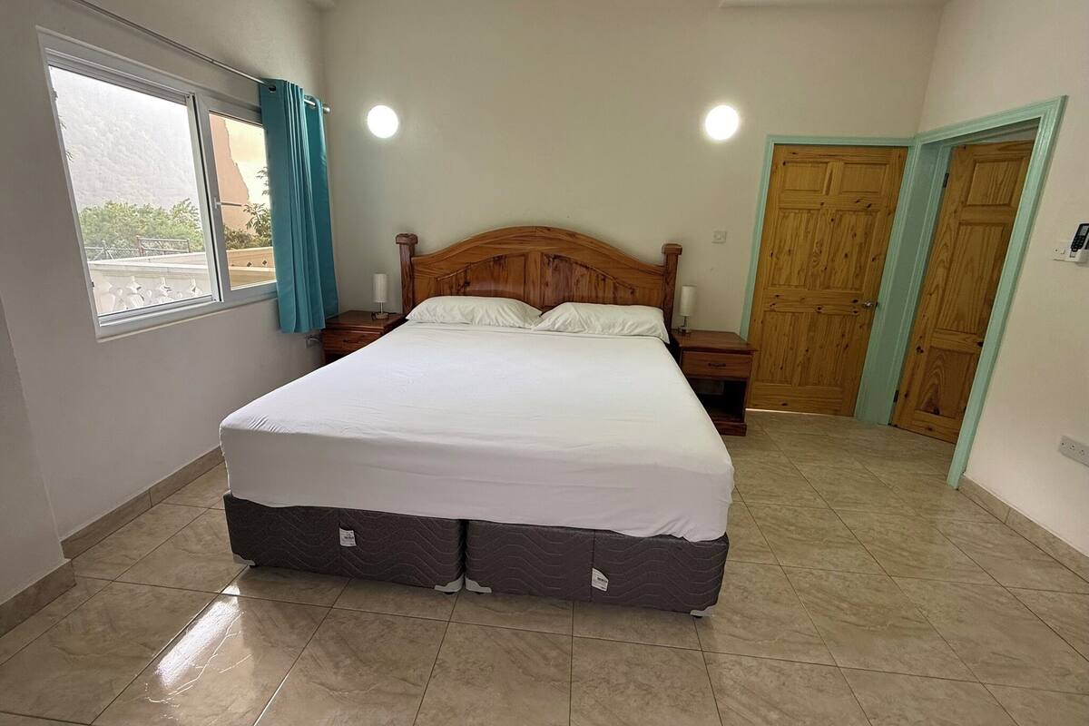 Ein Schlafzimmer mit einem großen Bett, einem Fenster mit Vorhängen, einem Nachttisch, einer Holztür und einem Fliesenboden.
