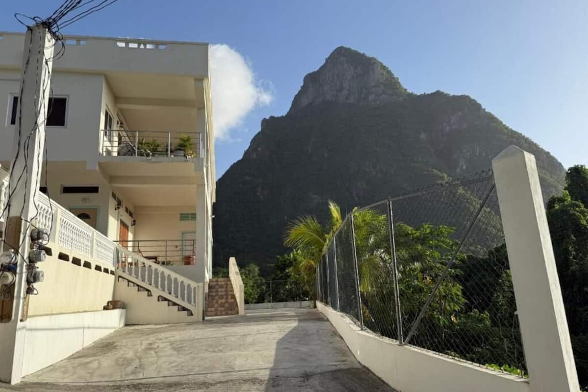 Ein Gebäude mit einer Treppe, die zu einem Balkon führt, einem Berg im Hintergrund und einem von einer Mauer umgebenen, begrünten Bereich.
