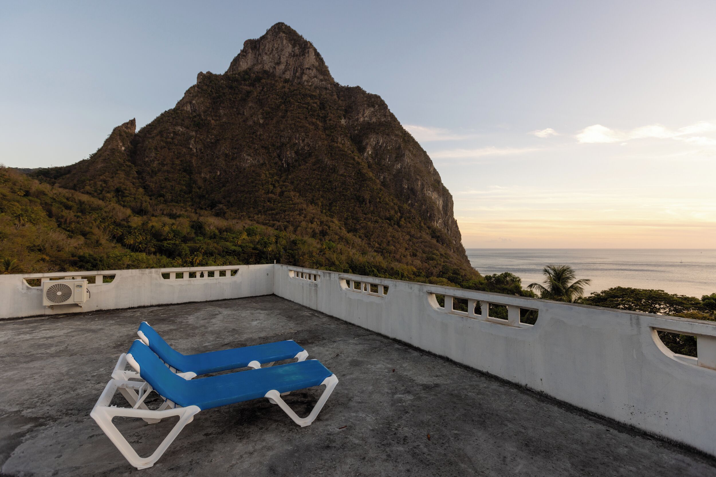 A rooftop terrace with a blue lounge chair, a white wall with ventilation, and a view of a mountain and ocean.
