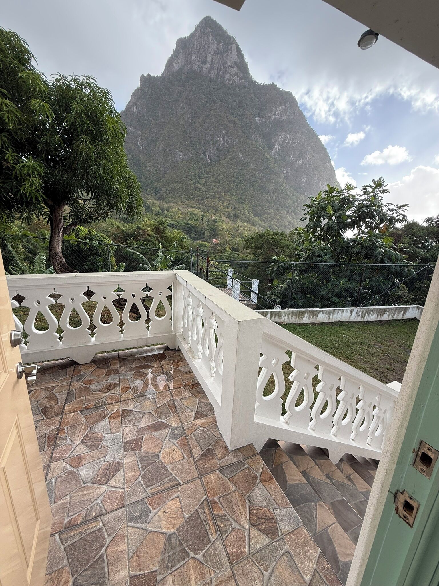 A balcony with a stone-tiled floor and white decorative railings, overlooking a mountainous landscape.