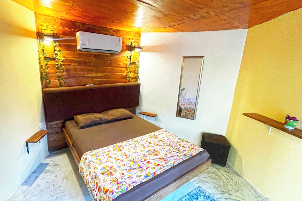 A bedroom with a bed, a nightstand, a wall-mounted air conditioner, a mirror, and a shelf with decorative items.