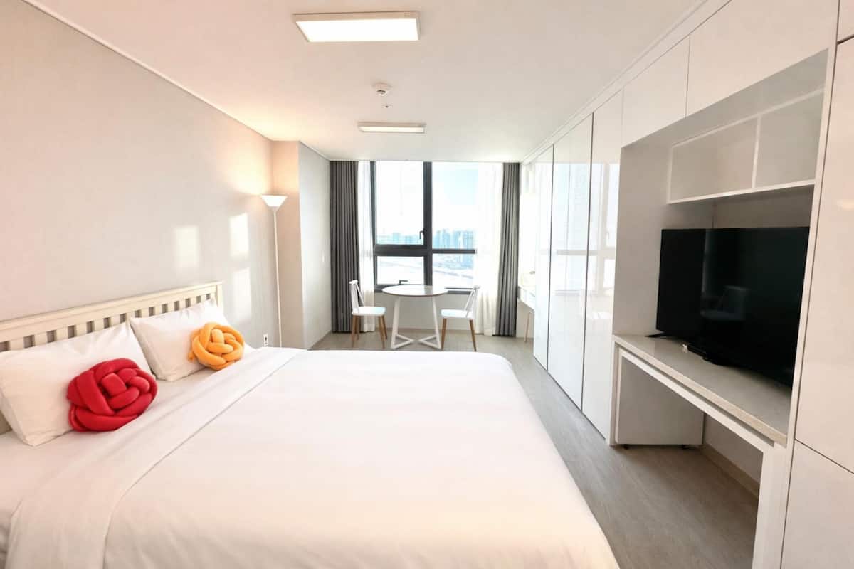 A hotel room with a bed, a TV, a desk, and a window with a view of buildings.
