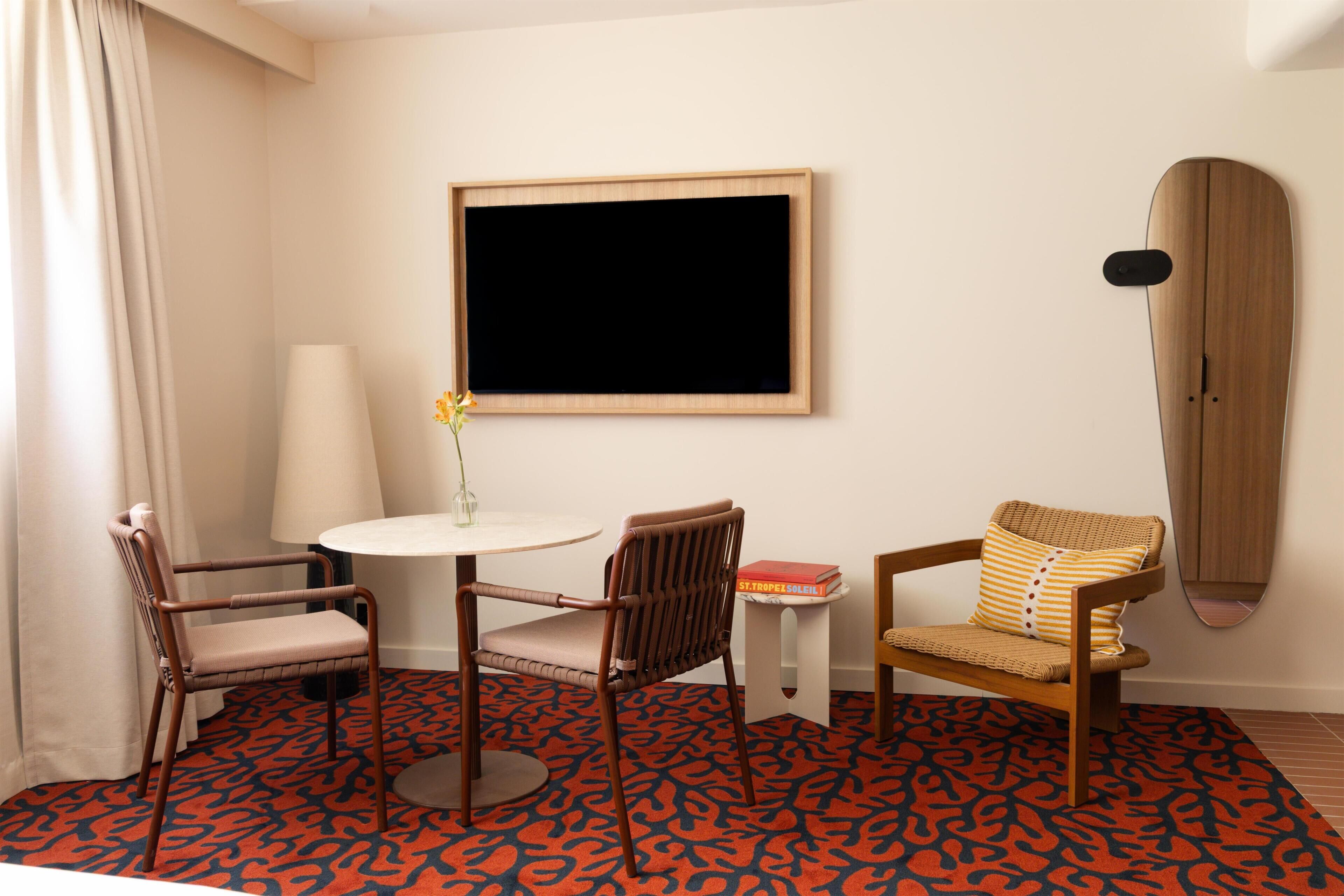 A hotel room with a round table, two chairs, a wall-mounted TV, a surfboard, and a patterned carpet.