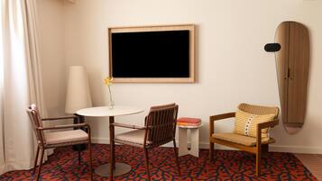A hotel room with a round table, two chairs, a wall-mounted TV, a surfboard, and a patterned carpet.