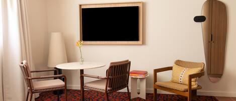 A hotel room with a round table, two chairs, a wall-mounted TV, a surfboard, and a patterned carpet.