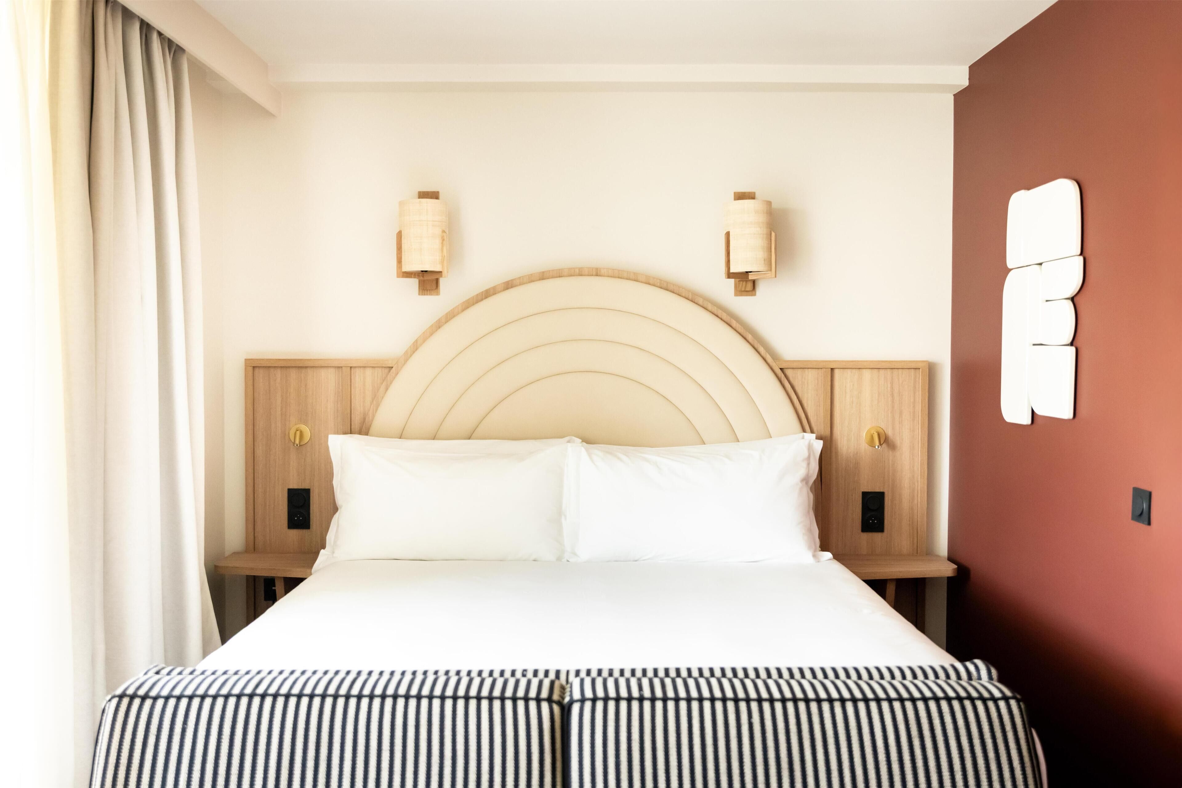 A hotel room with a bed, two bedside tables, a wooden headboard, and a wall-mounted light fixture.