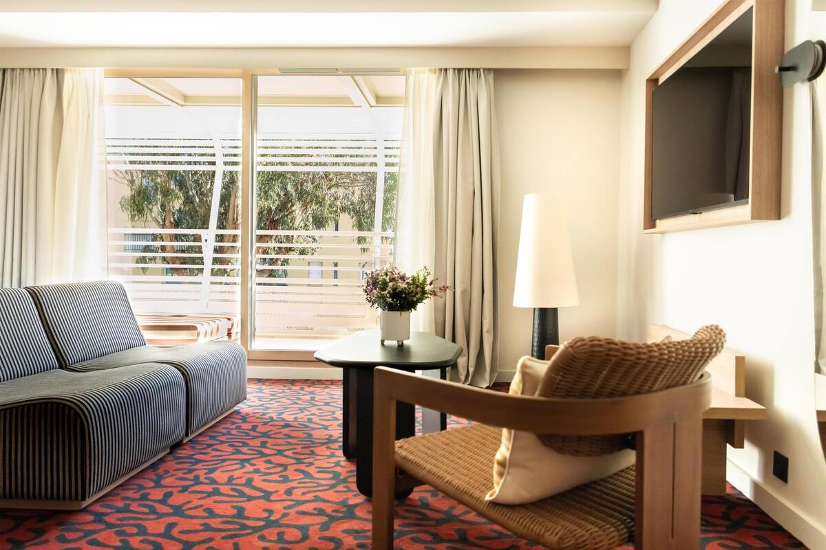Habitación de hotel con sofá, una silla, una mesita con un florero, una lámpara y un televisor en la pared.