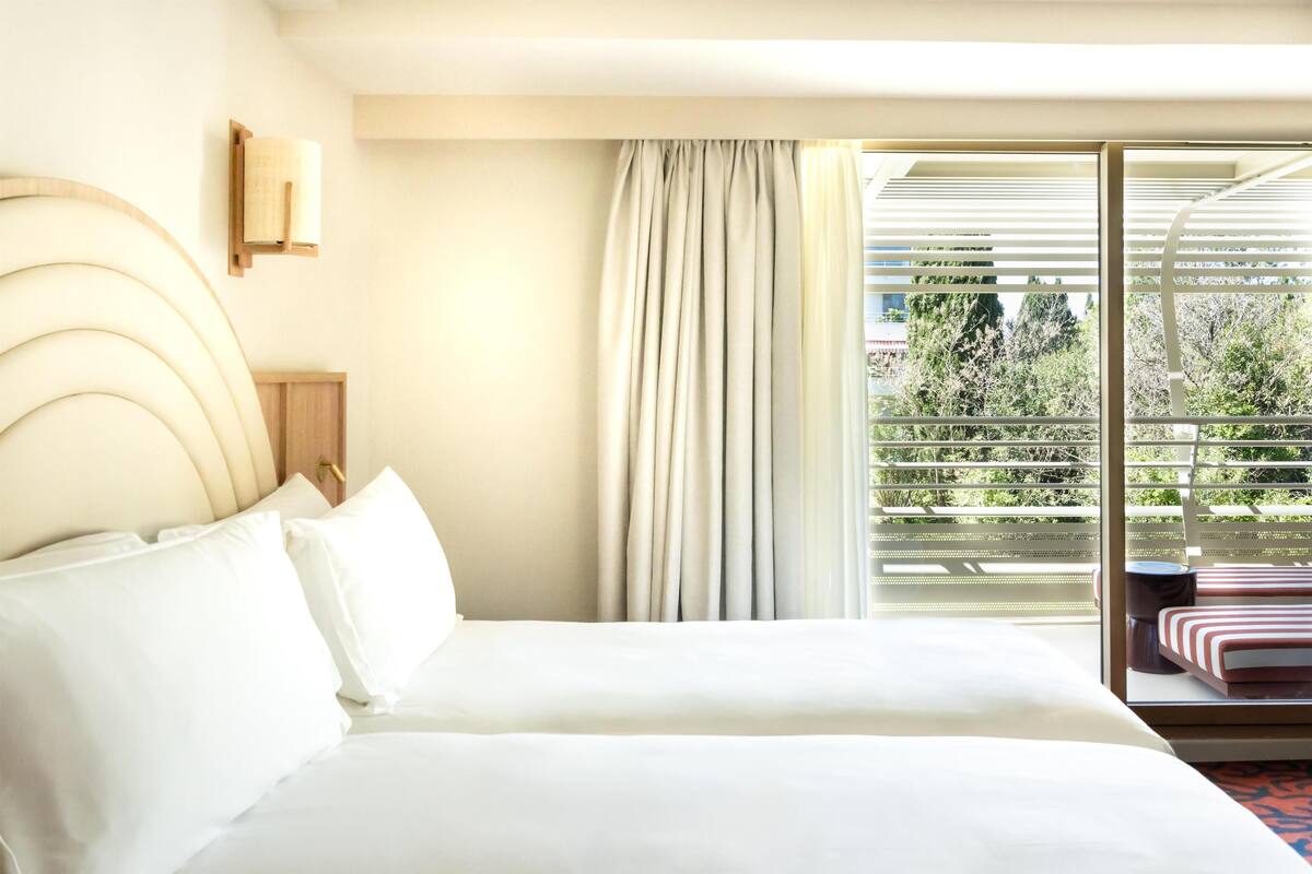 Una habitación de hotel con una cama, una mesita de noche, una lámpara, un ventanal con cortinas y vistas al exterior.