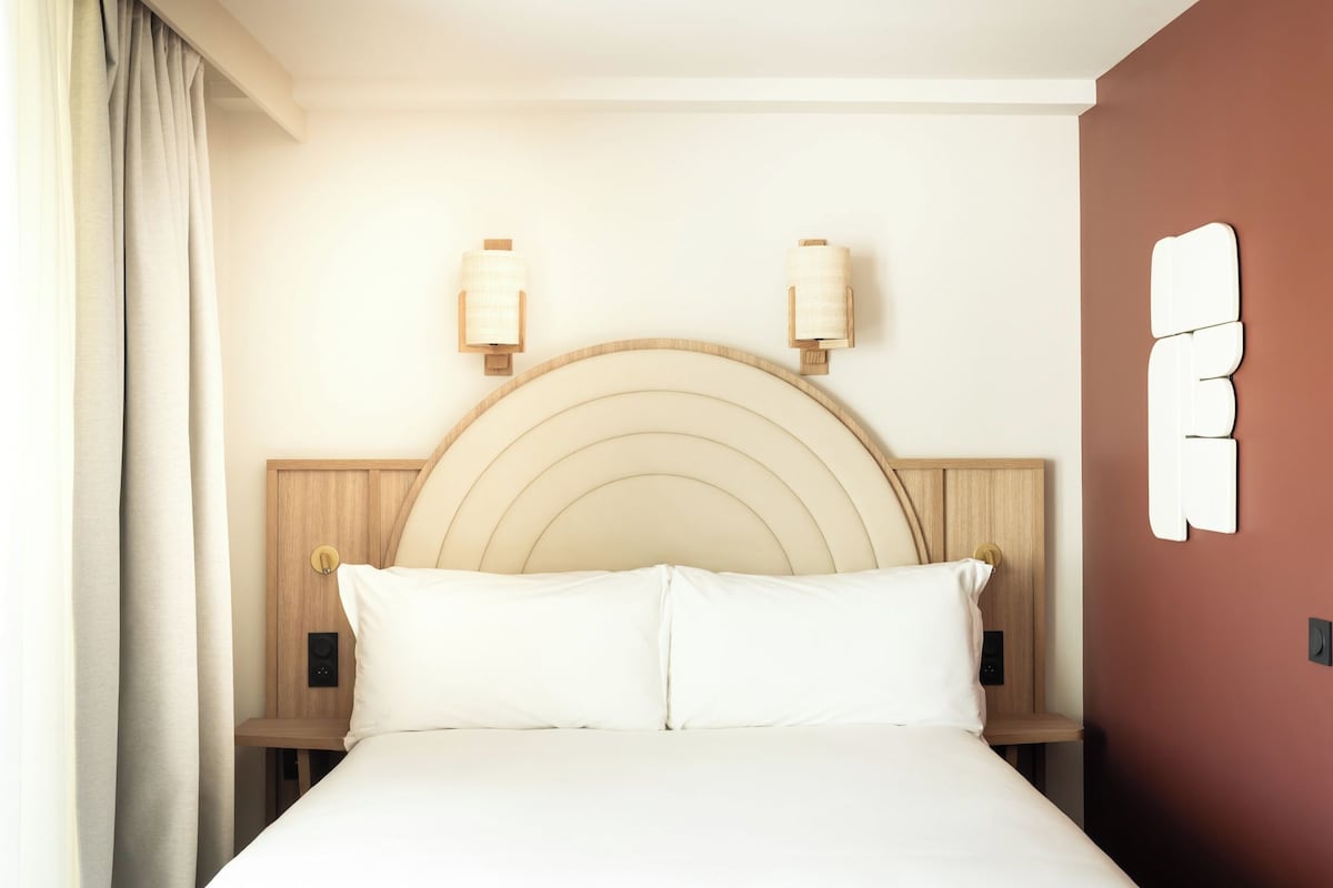 Habitación de hotel con cama, cabecero, mesitas de noche, lámparas fijadas a la pared y un reloj de pared.