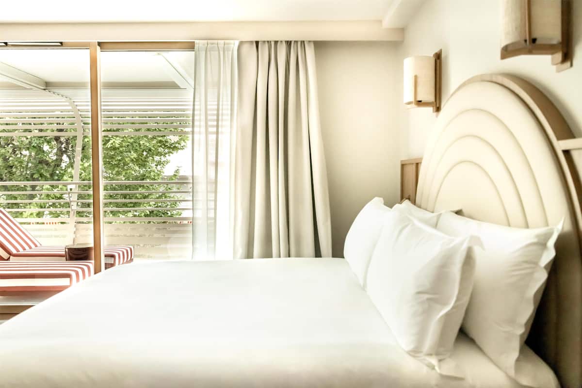 Habitación de hotel con cama, vista a zonas verdes y balcón con mobiliario a rayas.