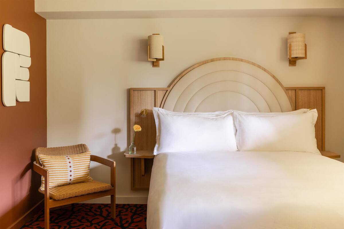 Una habitación de hotel con una cama, una silla, una mesita de noche y apliques de pared.