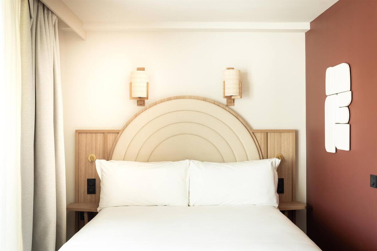 Una habitación de hotel con una cama, dos mesitas de noche, un cabecero e iluminación en la pared.