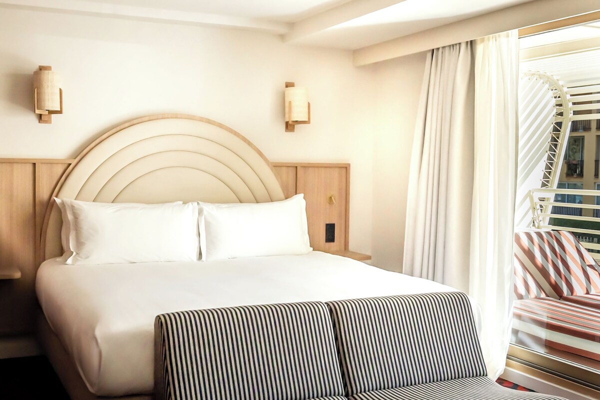 Habitación de hotel con una cama grande, cabecera de madera, dos lámparas fijadas a la pared y vistas a un balcón con mobiliario a rayas.