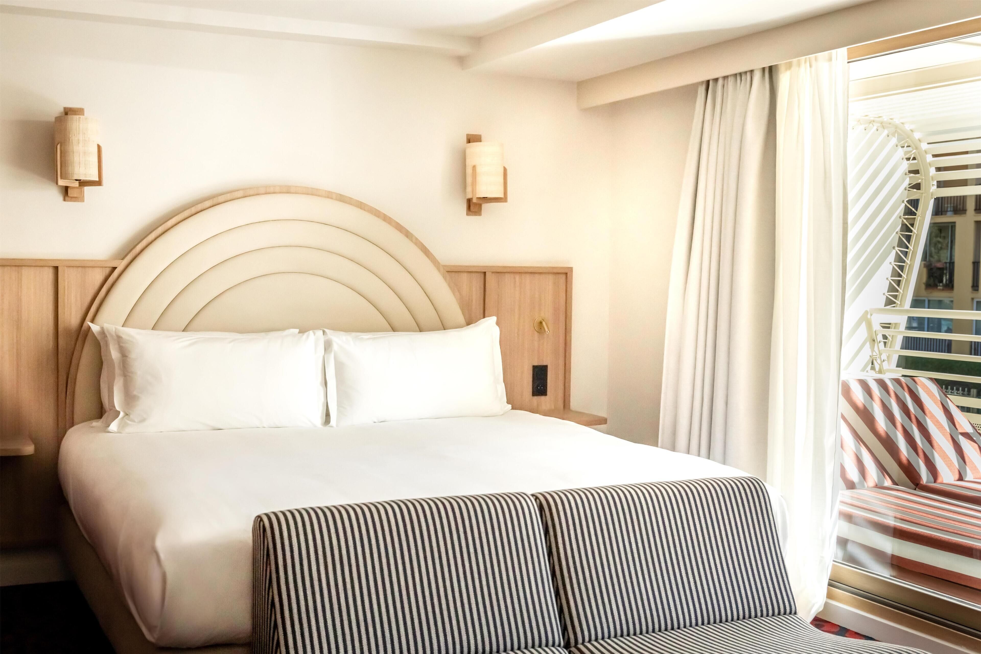 Una habitación de hotel con una cama grande, un cabecero de madera, dos apliques de pared, un ventanal con cortinas y un sofá a rayas.