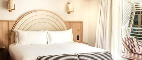 A hotel room with a large bed, a wooden headboard, two wall-mounted light fixtures, a window with curtains, and a striped sofa.