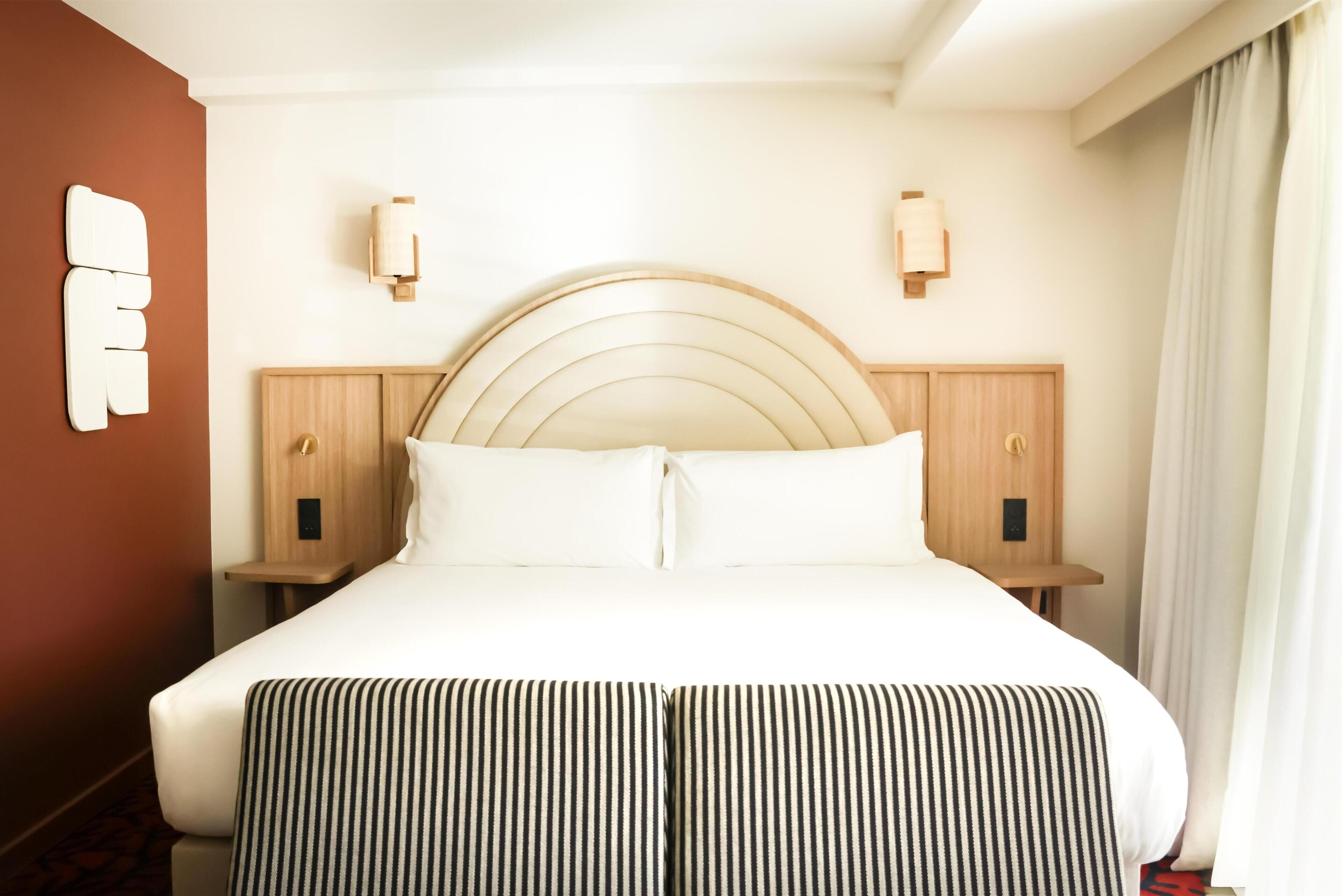 A hotel room with a large bed, two bedside tables, a wooden headboard, and a wall-mounted light fixture.