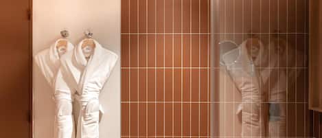 Un baño con espejo, lavamanos y dos albornoces blancos colgados en ganchos.