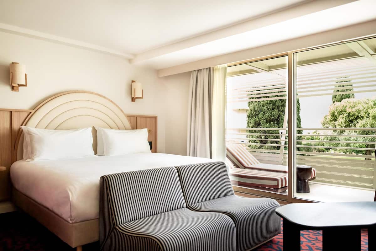 Una habitación de hotel con una cama, un sofá a rayas, una mesita y un ventanal con vistas a la vegetación.