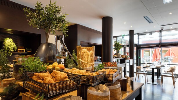 A breakfast buffet with various pastries and bread, a large potted plant, and a seating area with tables and chairs.