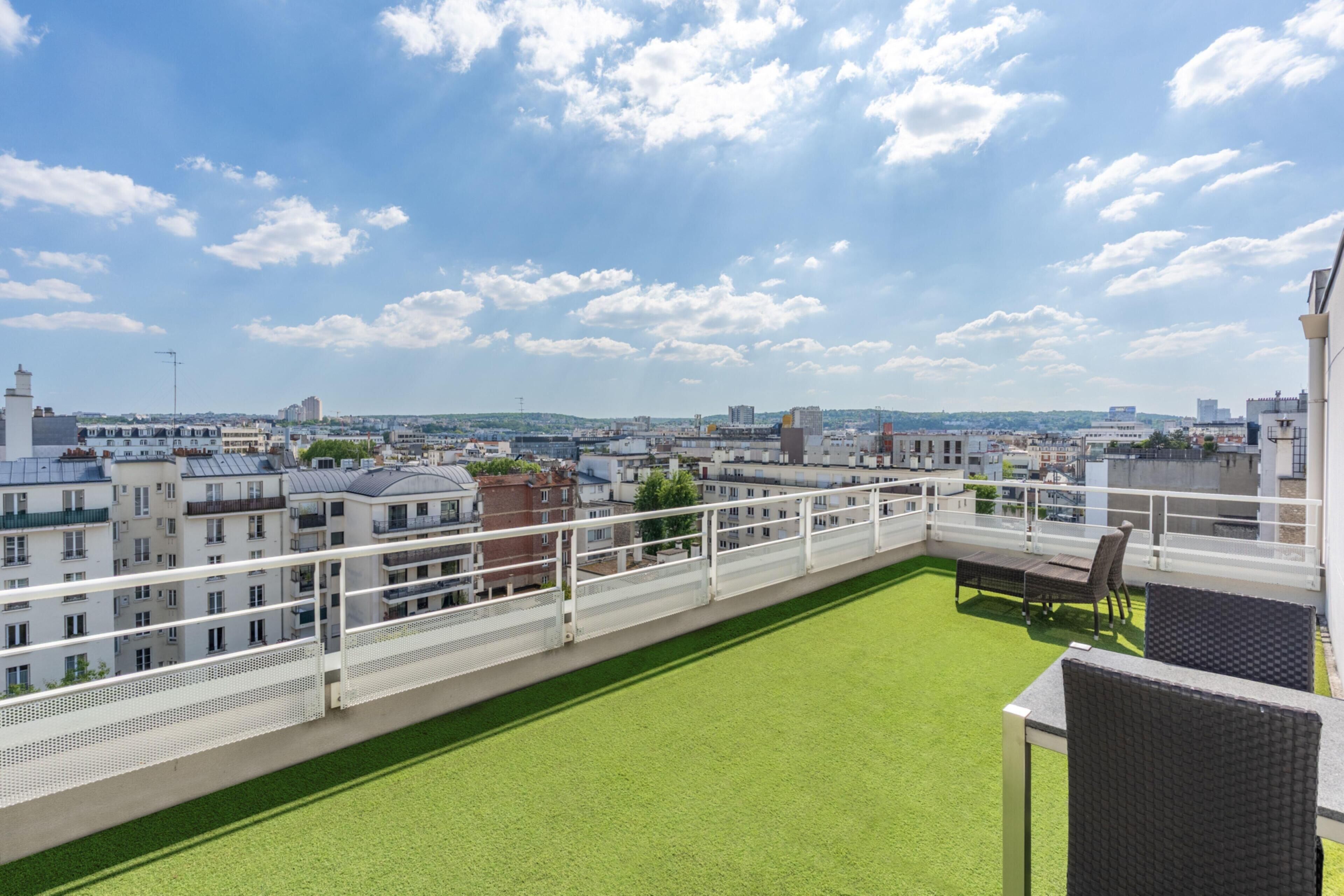 Une terrasse sur le toit offrant une vue sur le paysage urbain, avec une balustrade blanche et une pelouse verdoyante.