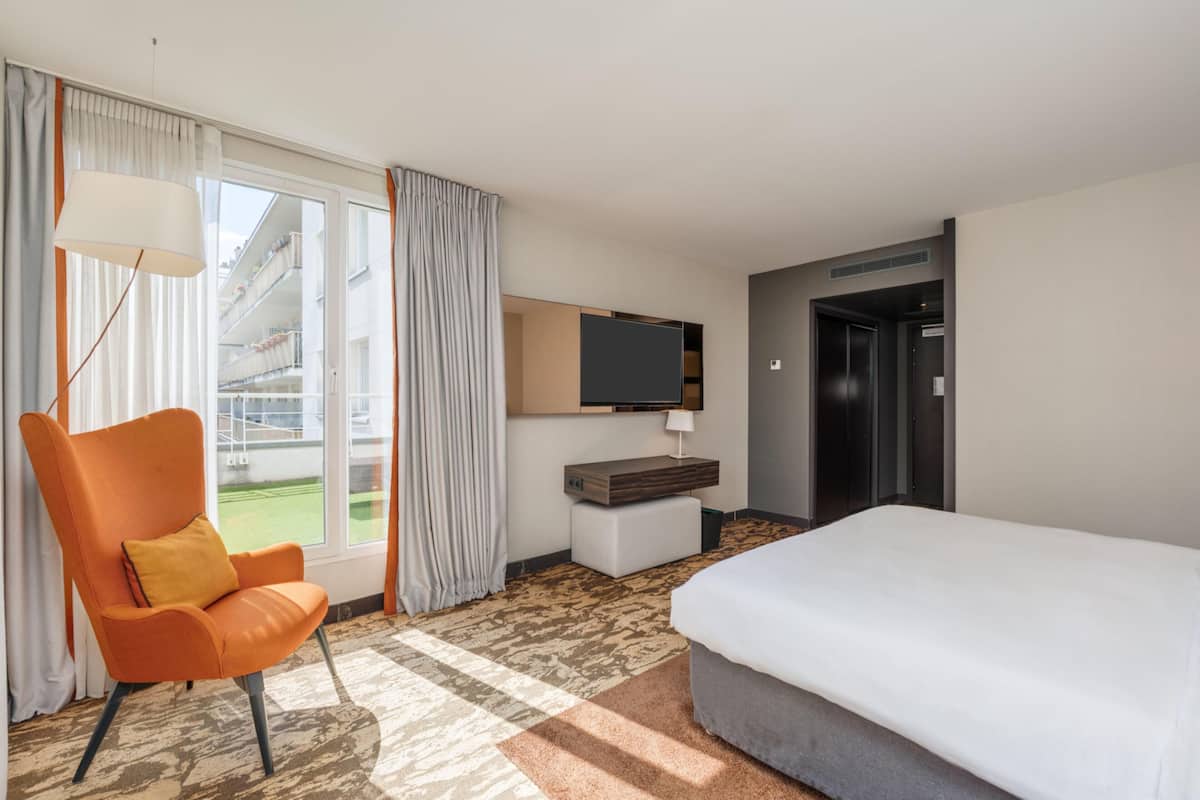 Une chambre d’hôtel avec un lit, une chaise, une télévision, un bureau et une vue sur un bâtiment à l’extérieur.