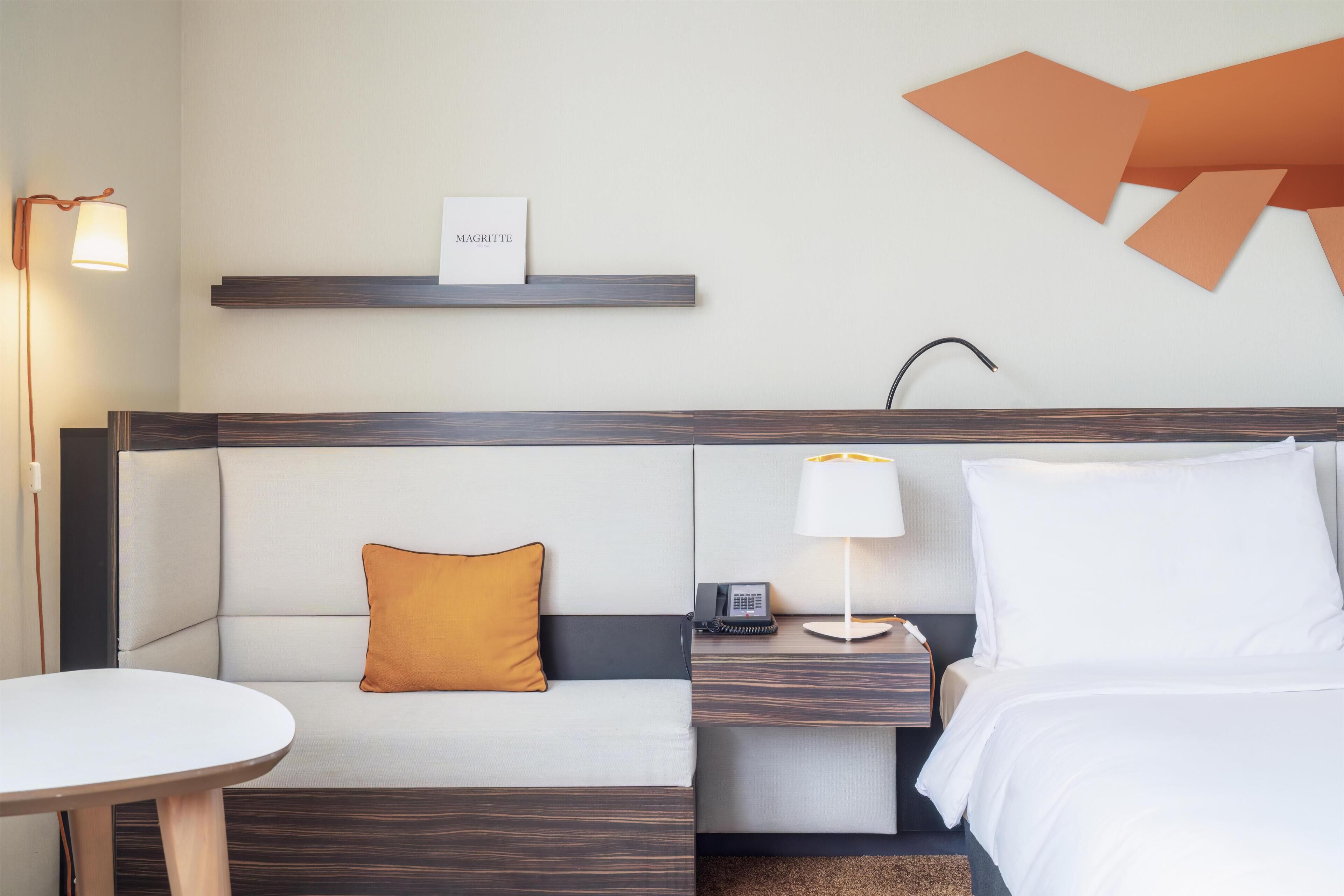 A hotel room with a bed, a bedside table with a lamp, a phone, a small shelf with a sign, a sofa with a pillow, and a round table.