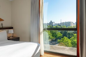 Habitación de hotel con un ventanal grande, una cama, una mesita de noche con lámpara y vistas a la ciudad.