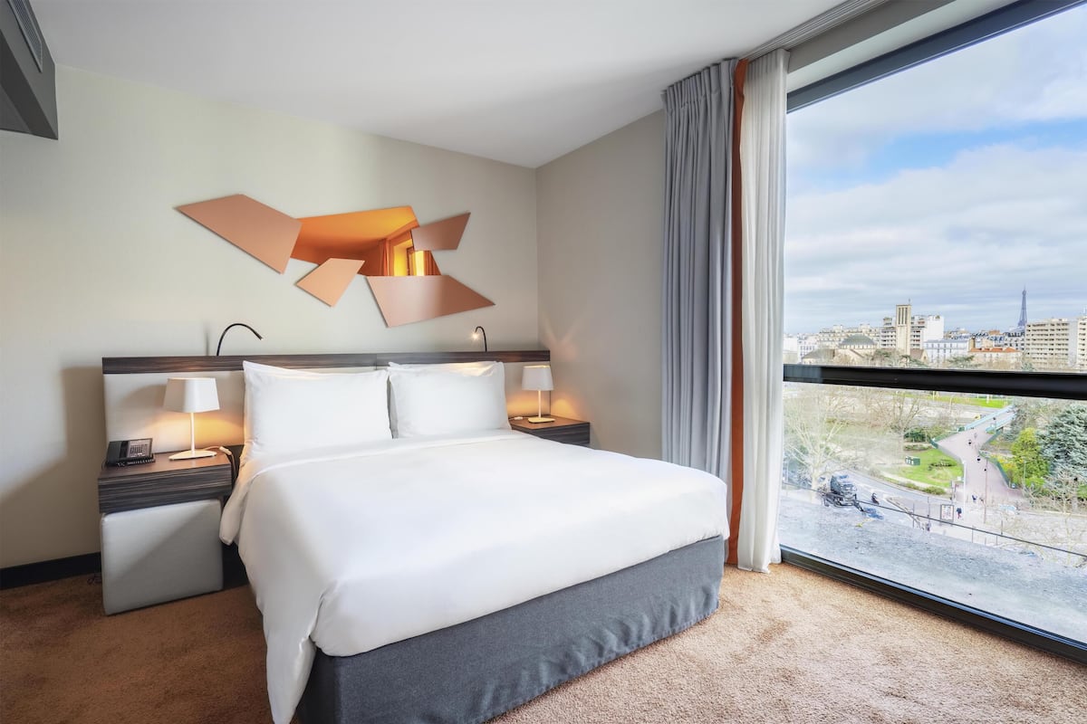 Une chambre d’hôtel avec un grand lit, des tables de chevet avec des lampes, une vue sur la ville par une grande fenêtre et une œuvre d’art moderne accrochée au mur.