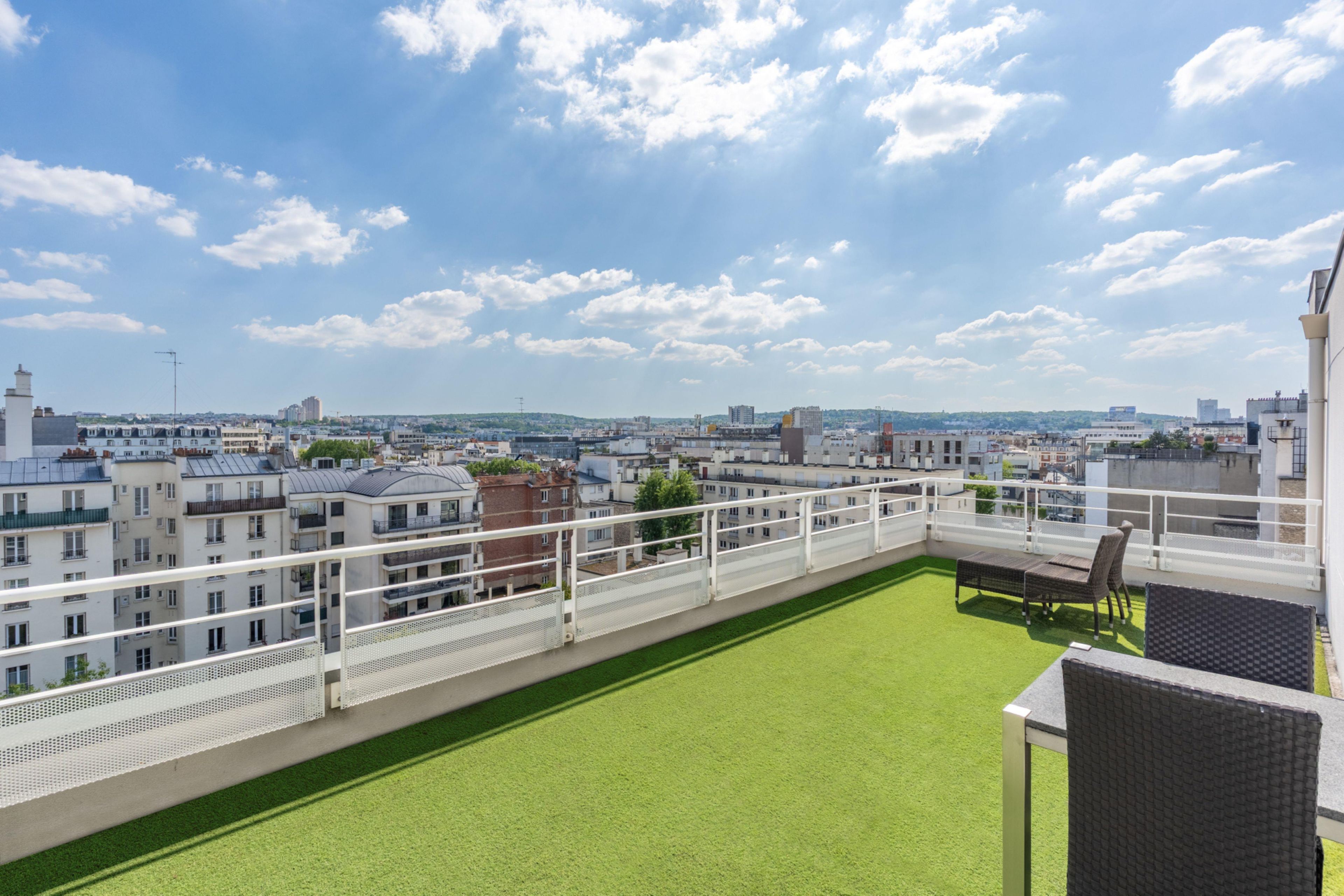 Una terraza en la azotea con césped verde y barandilla blanca, con vistas a un paisaje urbano con diversos edificios.