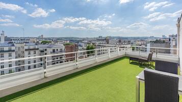 Una terraza en la azotea con césped verde y barandilla blanca, con vistas a un paisaje urbano con diversos edificios.