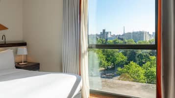 Ein Hotelzimmer mit großem Fenster, einem Bett, einem Nachttisch mit Lampe und Blick auf eine Stadtlandschaft.