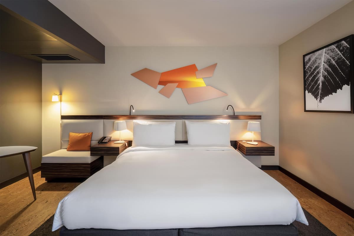 Une chambre d’hôtel avec un grand lit, des tables de chevet avec des lampes, une œuvre d’art abstraite accrochée au mur et une composition de feuilles encadrée sur le mur.