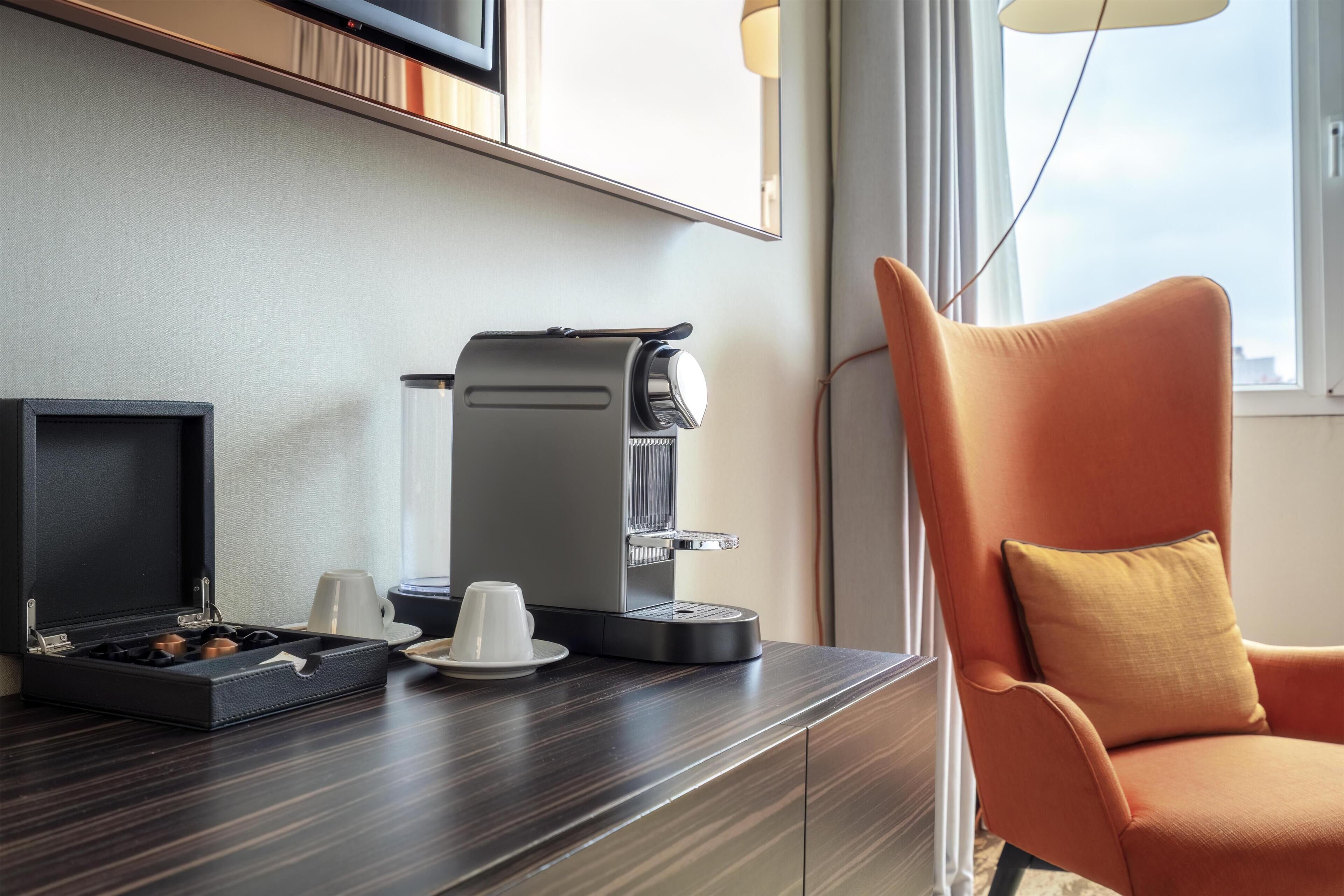 Ein modernes Hotelzimmer mit Kaffeemaschine, einem kleinen Tisch mit Tasse und Untertasse sowie einem gemütlichen orangefarbenen Sessel.