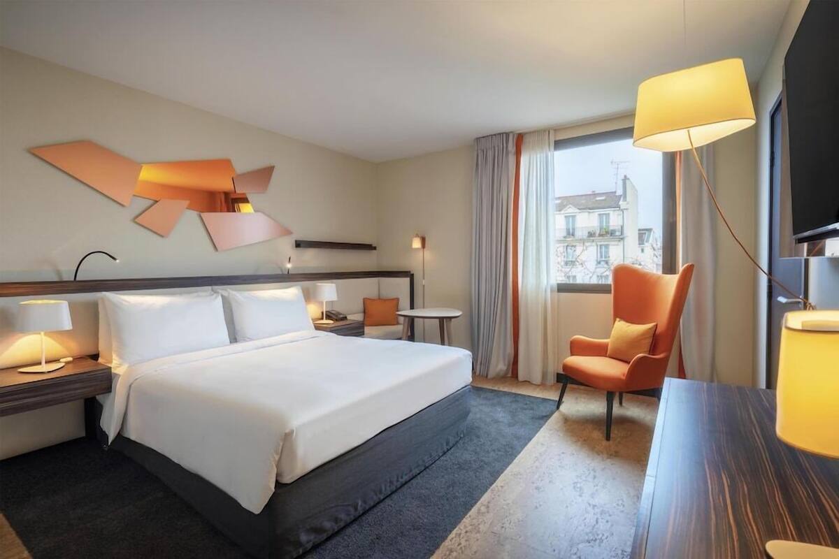 Une chambre d’hôtel avec un grand lit, un bureau, une chaise, une lampe et une vue sur les bâtiments à travers la fenêtre.