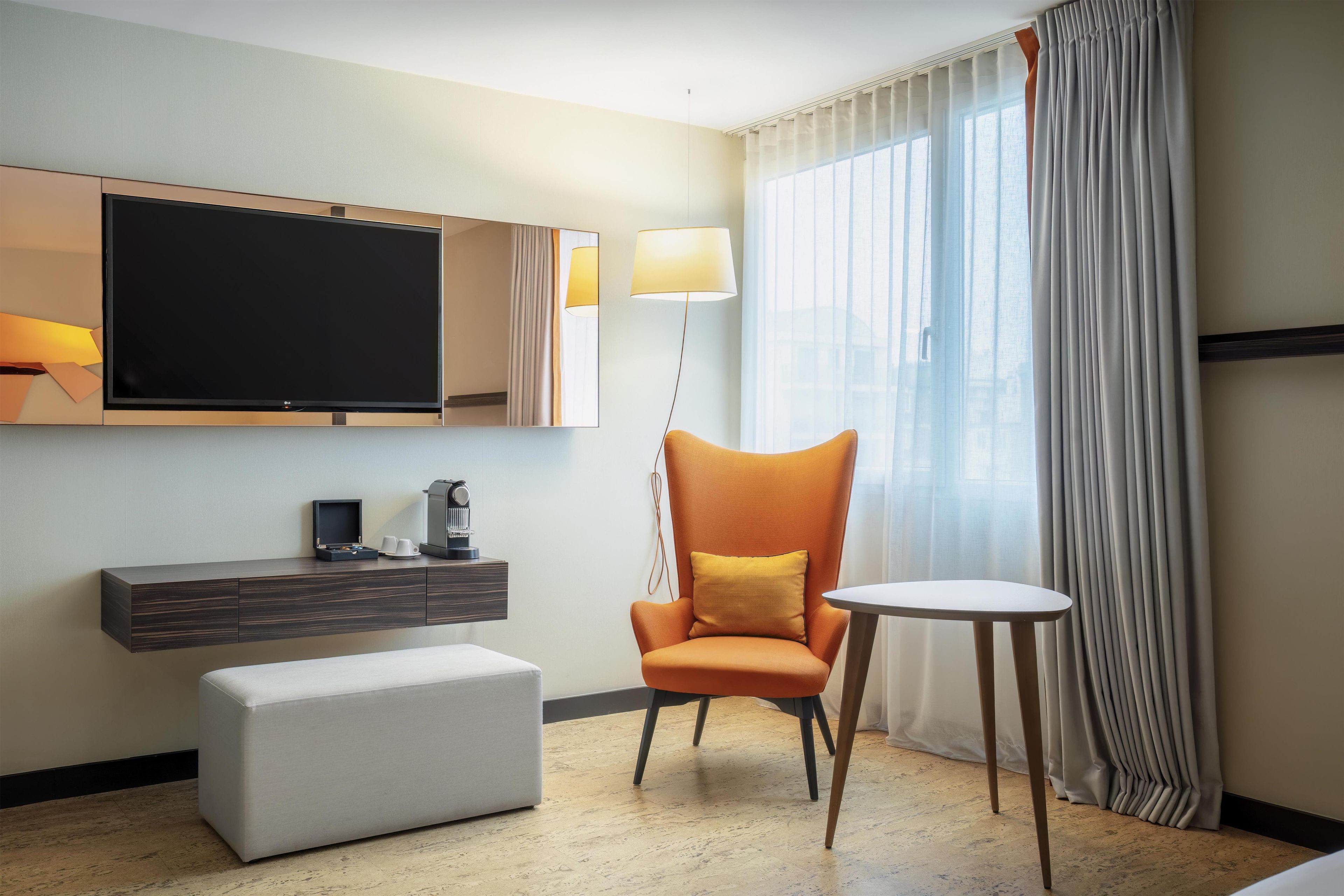 Une chambre d’hôtel équipée d’un téléviseur à écran plat, d’une machine à café, d’un canapé, d’un fauteuil, d’une petite table et d’une grande fenêtre avec des rideaux.