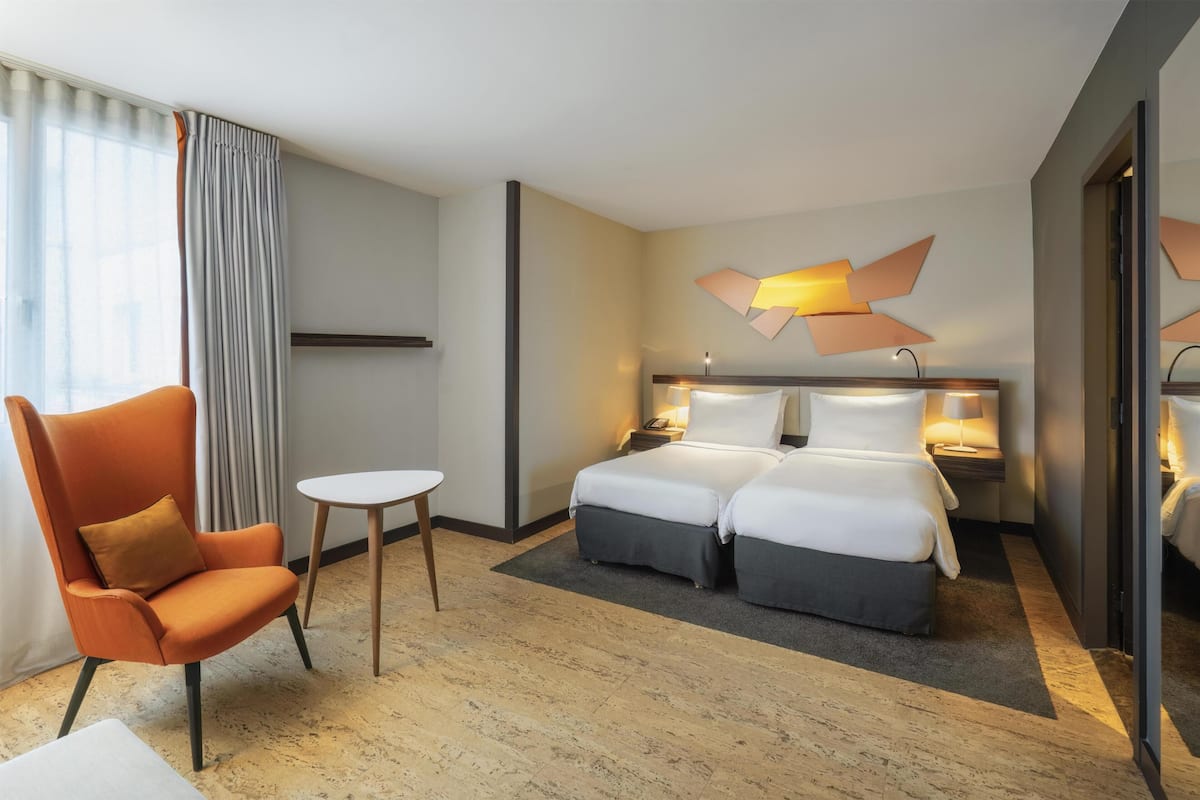 Une chambre d’hôtel comprenant un lit, deux tables de chevet, une chaise, une petite table et une grande fenêtre avec des rideaux.