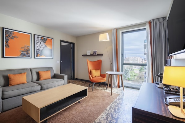 Una habitación de hotel moderna con sofá, sillón, mesita pequeña, escritorio y ventana grande.