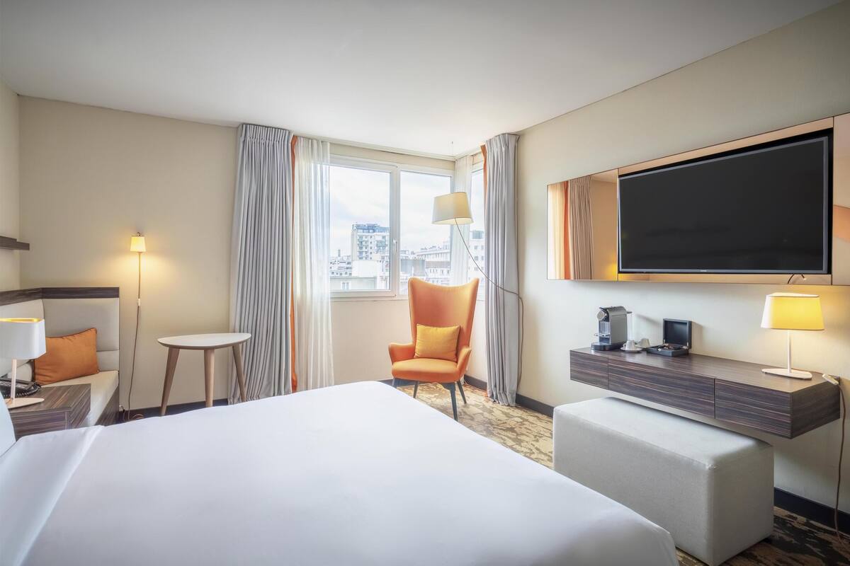 Une chambre d’hôtel avec un grand lit, un bureau, une chaise, une télévision et une fenêtre avec des rideaux.