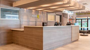 A modern hotel reception desk with a 'HOME 2' sign, a minibar, and a 7-Eleven kiosk.