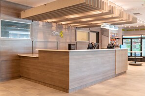 Un mostrador de recepción moderno de hotel con un cartel de 'HOME 2', una nevera y un quiosco de 7-Eleven.