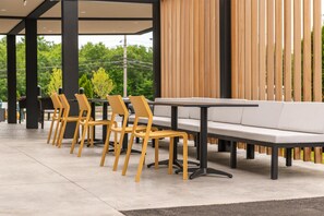 Un área de estar moderna al aire libre con una fila de sillas amarillas y un sofá blanco, suelo de hormigón y un muro de listones de madera al fondo.