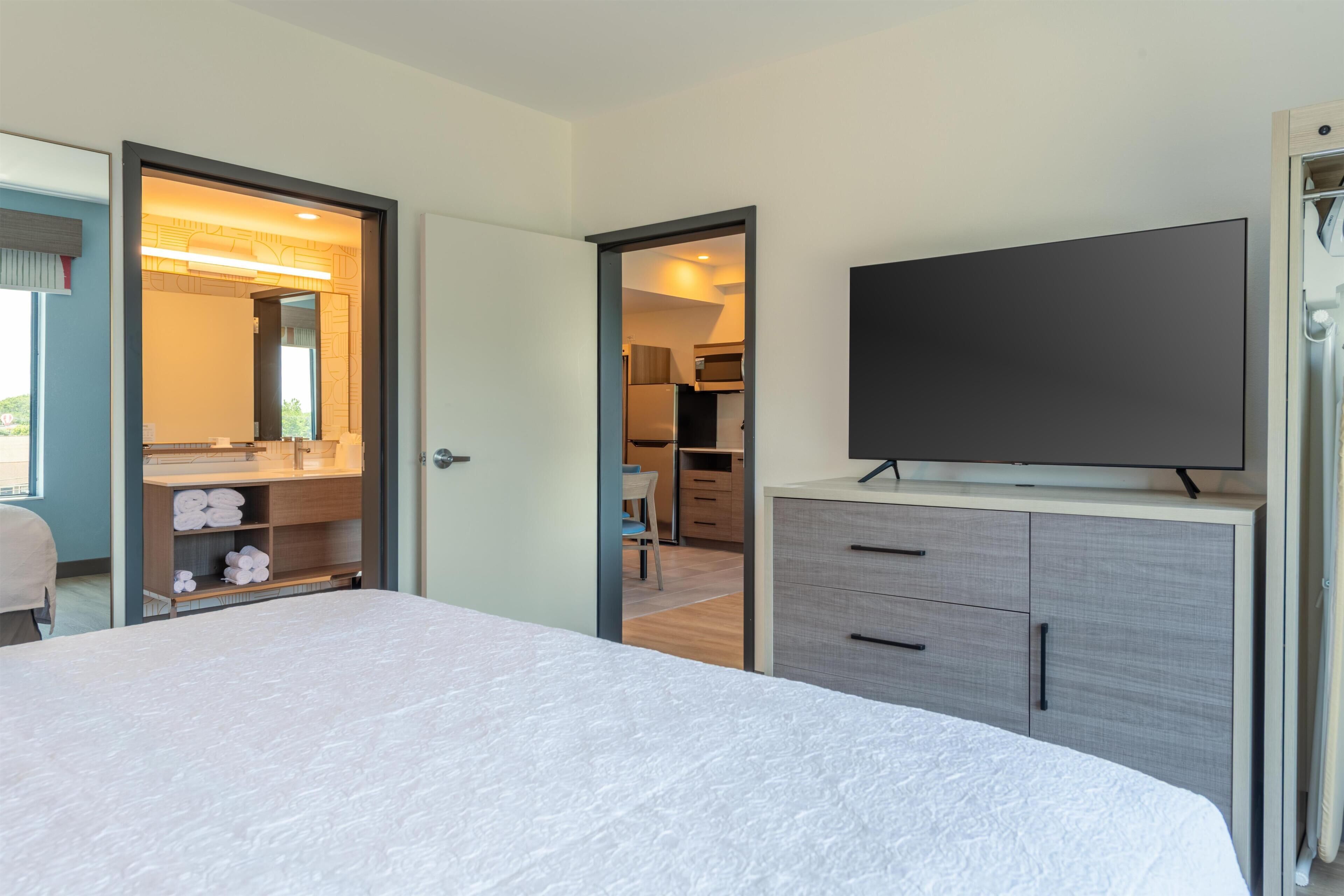Habitación de hotel con cama, televisor, armario y baño visible a través de una puerta abierta.
