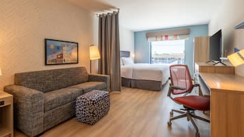A hotel room with a sofa, a patterned ottoman, a desk with a chair, a bed, and a window with a view.