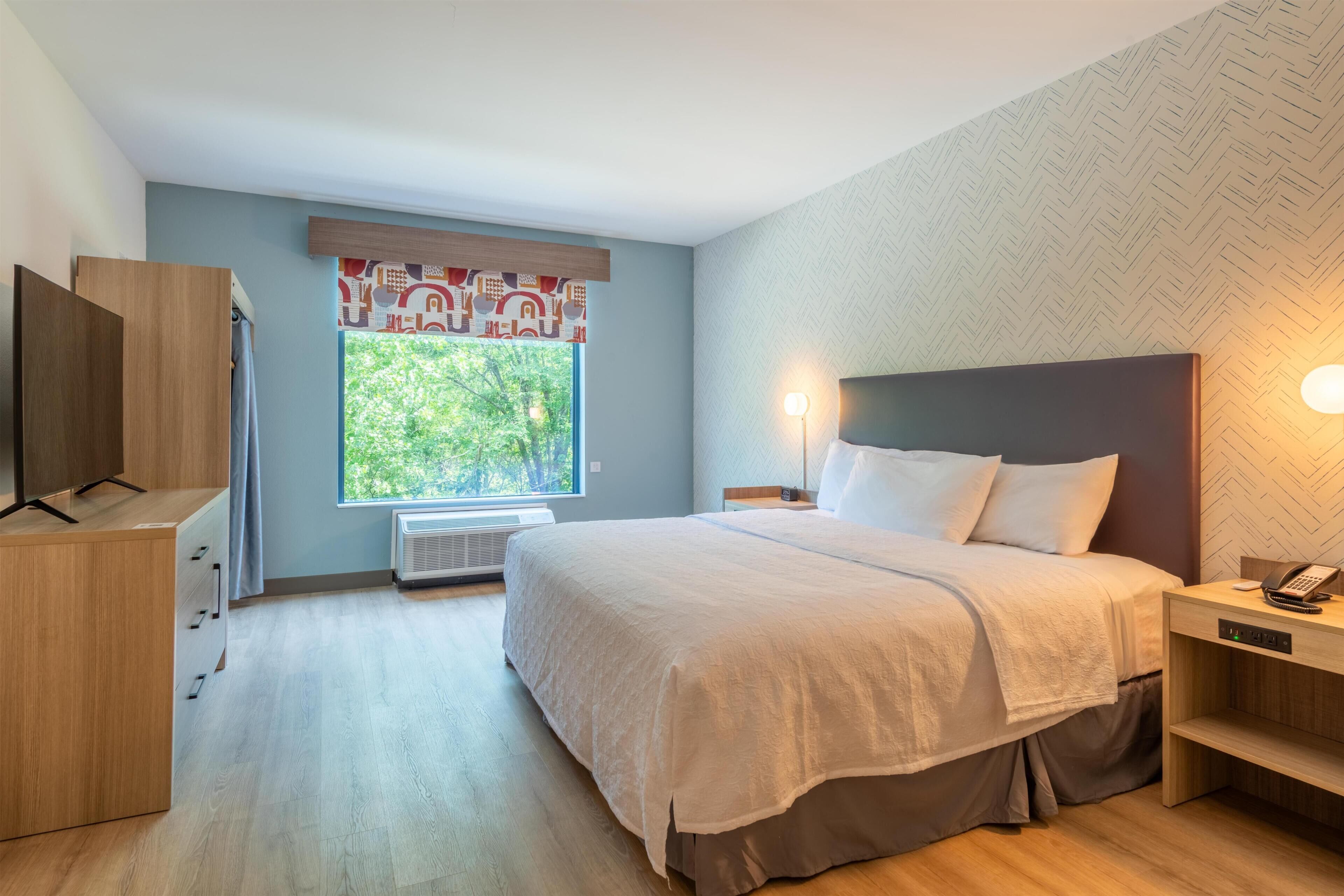 Een hotelkamer met een bed, nachtkastje, lamp en uitzicht op bomen door het raam.